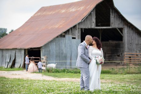 A man and woman kissing in front of an old barn.