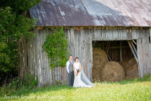 A bride and groom standing in front of an old barn.