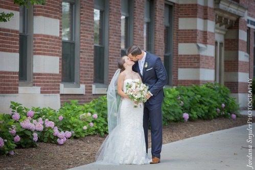 A bride and groom kissing on the side of a sidewalk.