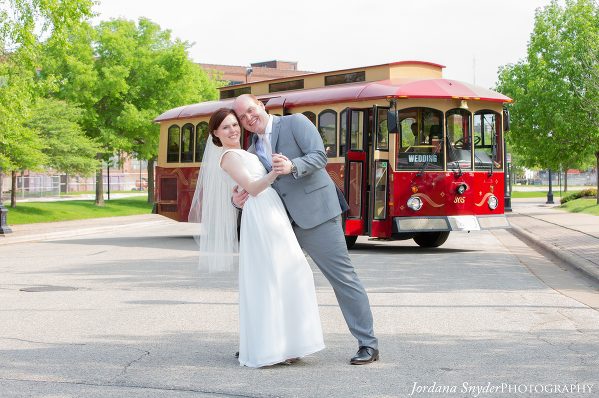 A bride and groom pose for the camera in front of a trolley.