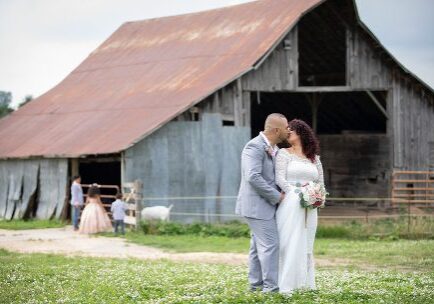 A man and woman kissing in front of an old barn.