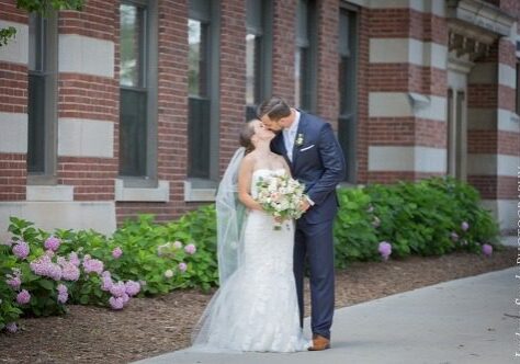 A bride and groom kissing on the side of a sidewalk.
