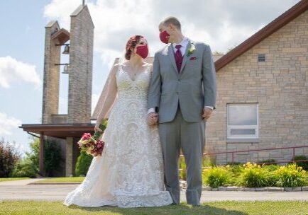 A bride and groom stand in front of their home.