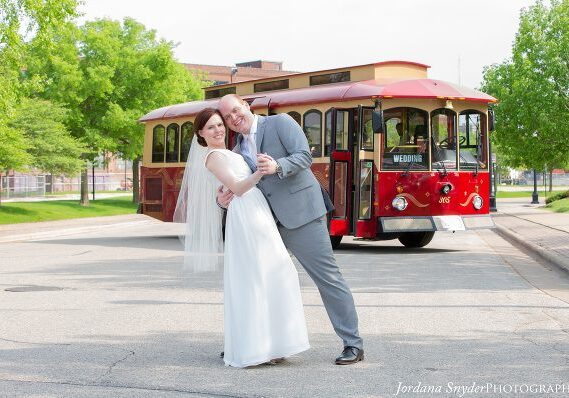 A bride and groom pose for the camera in front of a trolley.